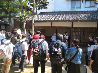 瓦屋根のある建物の下で話をする男性2人とその方向を向いて話を聞くリュックを背負った学生達の後ろ姿が写っている写真