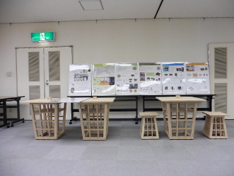 室内の壁側に立てかけられた6枚の展示物と、その前に格子状の脚をした机3台と椅子2脚が置いてある写真