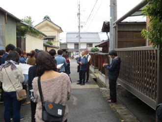 住宅街の道路で右側に黒い服やスーツを着た男性が立っており、その人達を見るように左側に信州大学工学部建築学科の生徒たちが画版を持つ等して立っている様子の写真