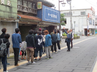 石畳の道路の左側に赤い屋根や青い看板が設置された建物を指さすスーツ姿の男性とまばらに並び目の前の建物を見る信州大学工学部建築学科の生徒達の「街区の再生」授業の様子の写真