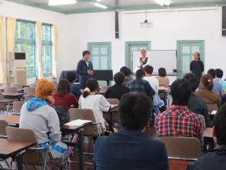 机に資料を置き座っている学生達と、ホワイトボードの前に白シャツ灰色ベストを着た男性が話している授業の様子の写真