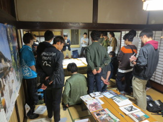 和室に資料が置いてある長机があり、奥の和室を向くように1人は座りそれ以外の学生達は立って見学している様子の写真