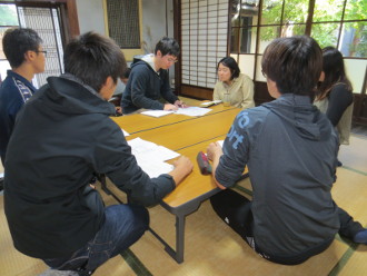 縁側のある和室に机を置きそれを囲う様に座り、資料を見て話をしている学生達の写真
