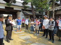 旧上高井郡役所の飛び石のある庭で、長野工業高等専門学校の建築設計製図の生徒たちに向かって灰色のスーツを着た男性が資料を持ち話をしている授業の様子の写真