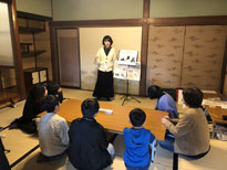 和室で長机半分を囲う様に座る大人や子ども達の前で資料をスタンドにたてかけ、話をする黒のロングスカートに白のカーディガンを着た女性の写真