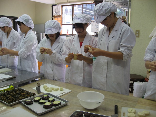 テーブルにあんや包丁、ボールなどが置かれ、給食帽や調理衣を着た5人の女性が生地を薄く伸ばしておやきの皮を作ろうとしている様子の写真