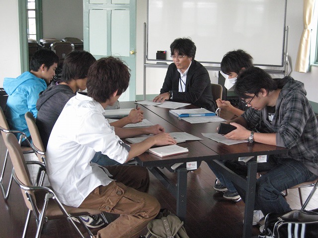 長机に資料を置き、対面する形で男子生徒が3名と2名、その左隣に黒いジャケットを着た教員男性1人が座り、授業をしている写真