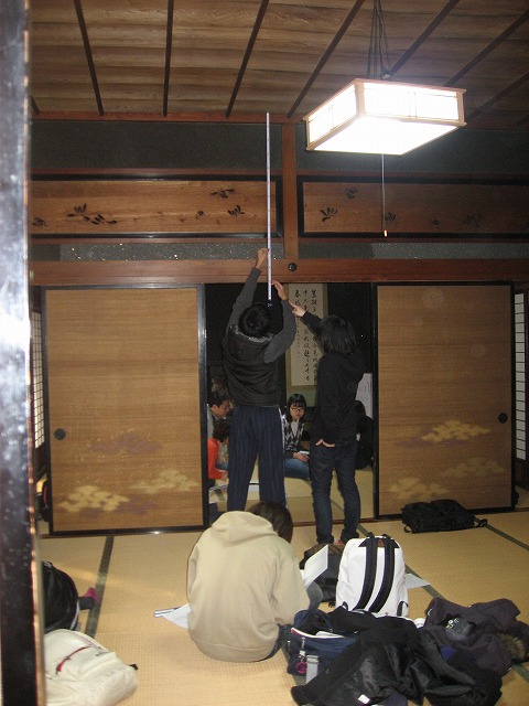 畳と襖がある趣きのある民家の和室の中央に立ち、メジャーを使い天井の高さを測っている2人と座って資料を見ている生徒たちの実習の様子の写真
