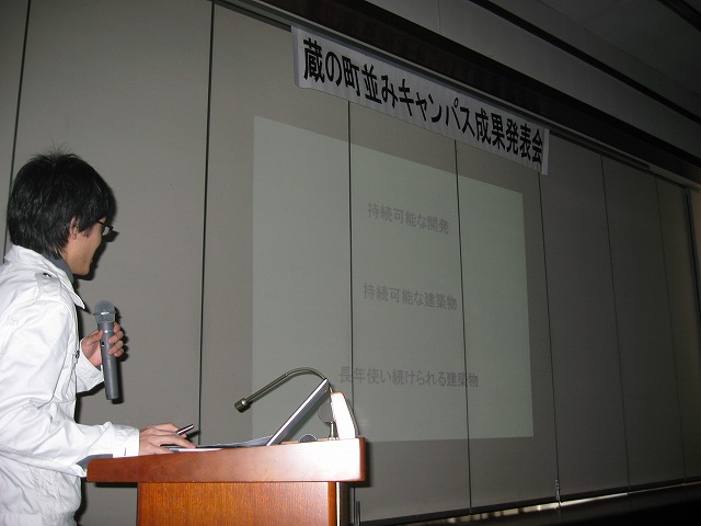 「蔵の町並みキャンパス成果発表会」と書かれた貼り紙の下のスクリーンに映し出された映像を見ながらマイクで話をしている男性の写真