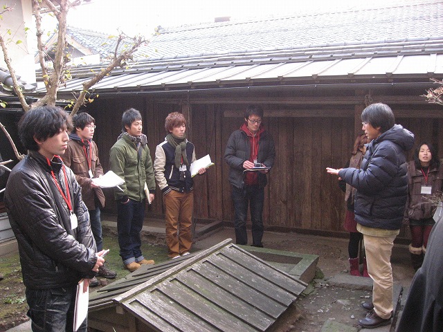 屋根が低い木造の建物の横に、中央に屋根らしき物があり、それを囲う様に左側に男子生徒、右側に先生が立って説明をしている様子の写真