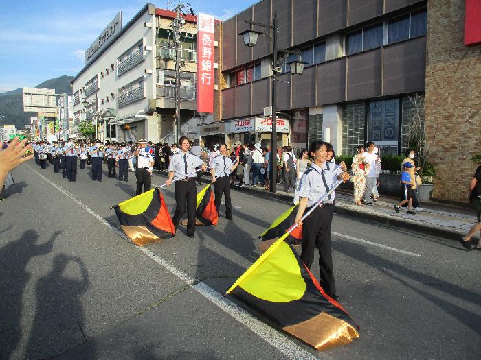女性消防団員4名で、黄色、黒、赤、金の4色で彩られたフラッグを持ち、お祭りで音楽隊とともにパレードする様子を写した写真