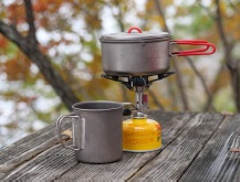 屋外の木製テーブルの上でガスバーナーコンロで鍋で湯を沸かしている。そばには鍋より一回り小さいマグカップが置かれている。