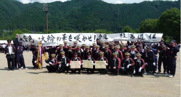 1列目の中央の人3名が賞状、左端の人が優勝旗を持ちながら横断幕の下に並びながら記念撮影をしている消防団員の方たちの写真