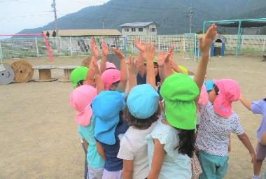 豊丘保育園の園庭で異年齢の園児が、指で一を作りみんなで集まっています。ピンク、黄緑、水色など異年齢の帽子が集まっています。