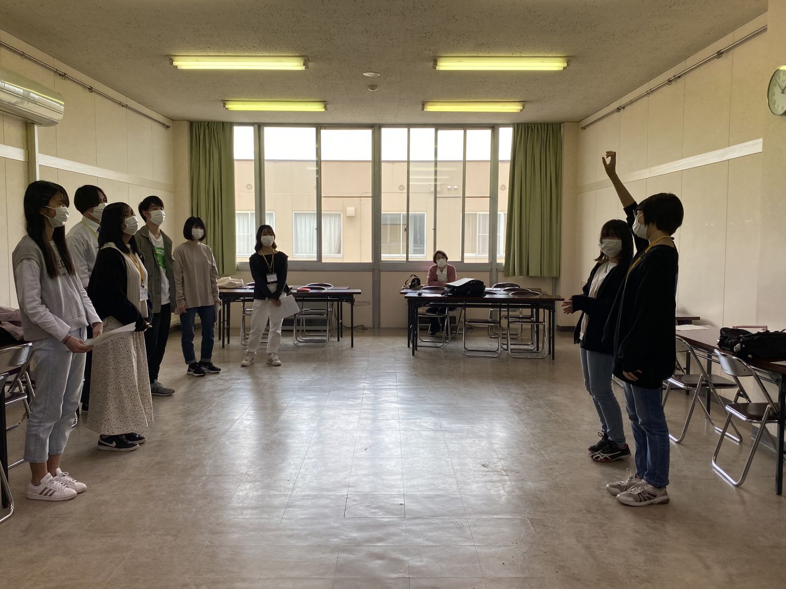 大きい窓がある室内で、参加者が向かい合って立ち、手前右側に立っている女性が手をあげている写真