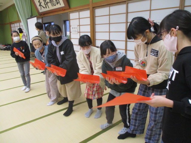 広い畳の上で、7人の女子児童が横一列に並び、手前にいる女子生徒の手の動きを見ながらオレンジの紙を二つ折りにして持っている様子の写真