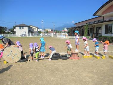広い園庭の中央に、木のはしご、木の板、添え木、タイヤなどを集めて作った自分たちのアスレチックに、園児たちが並んで遊んでいる様子。