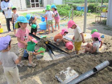 相之島保育園の広い砂場の中央に大きな砂のトンネルがあり、そのトンネルを囲んで園児たちが泥んこ遊びをしている風景です。トンネルから水を流したり、どろみずで遊んだりどろんこ遊びを楽しんでいます。