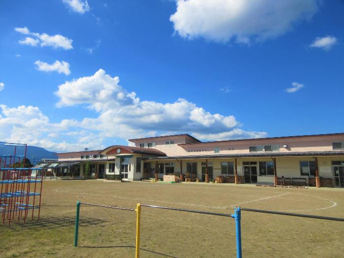 青空を背景に薄ピンク色の園舎がはえる相之島保育園の園舎写真。中央が玄関。Iの字の園舎の前には園庭が広がり、園庭の一角には鉄棒やジャングルジムがあります。