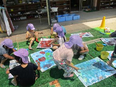 園庭の日陰のシートを敷いた場所に、園児たちが座り、それぞれ違った大きさの紙に青や赤や黄色の色水を使い絵を描いている様子。青でいっぱいの絵や手形がついた絵、赤でいっぱいの絵などいろいろな絵が描かれている。