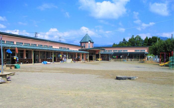 晴れた青空に日野保育園のピンクの外壁が生える写真。1階建ての園舎。エル字型の園舎の前に広い園庭。園舎の中央にある玄関の上は三角の屋根。