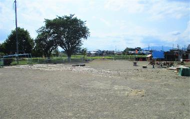 日野保育園の広々とした園庭を園舎側から撮影した写真。正面には大きな木、園庭のまわりにはうんていやアスレチックなどがある。