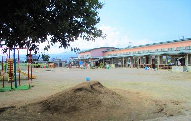 日野保育園園庭を玄関から撮影した写真。園庭を挟んで3歳以上児の園舎が見える。手前には園児が作った大きな土の山。