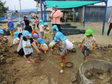 井上保育園の泥んこ遊びの様子。はだしの園児たちが園庭で泥の地面の感触を楽しみながら遊んでいる。泥団子を作っている子、スコップで遊んでいる子などの様子。