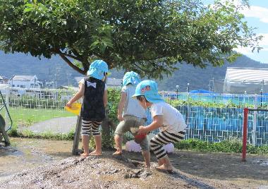 井上保育園の泥んこ遊びの様子。はだしの3人の園児が園庭で小高い土の山から水を流したりして泥の感触をたのしんでいる。