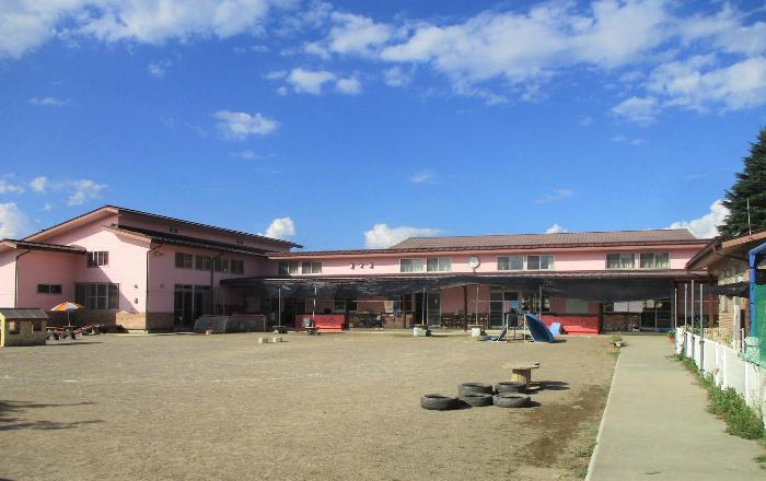 青空の下、ピンク色の外壁が映える井上保育園の園舎写真。1階建て。左側には片流れ屋根で園舎より高い建物のホール、園舎の前には園庭が広がります。