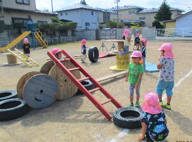 北旭ケ丘保育園の広い園庭の真ん中に、木のはしごや木の平均台、木のたる、タイヤなどを運び。園児がそれを組み合わせてアスレチックを作っている様子。