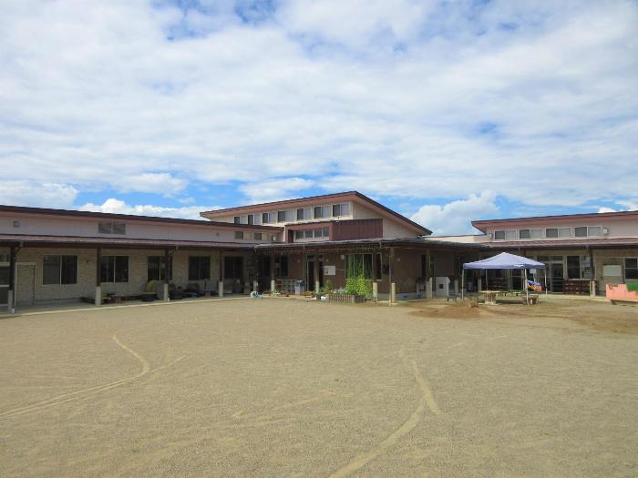 北旭ケ丘保育園園舎の写真。白い外壁の1階建ての建物。L字型の建物の中央に玄関、玄関の屋根は片流れ、園舎前には園庭が広がる。