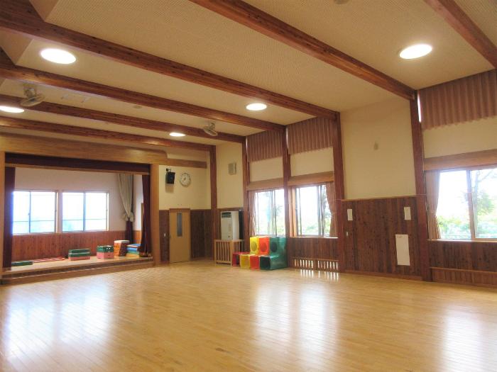 北旭ケ丘保育園ホールの写真。天井には木の梁があり、木の内装で作られ、温かみがあるホール。大き目の窓からは日差しが差し込み明るい。奥にはステージ、ホール内にはいろとりどりのブロックや跳び箱が置いてあります。