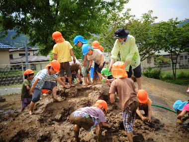 園庭の大きな砂山に水を流し、泥んこ遊びを楽しむ園児。全身を泥だらけにして砂山に登ったり、泥の感触を楽しんだり異年齢の児童が混ざって遊んでいる様子。