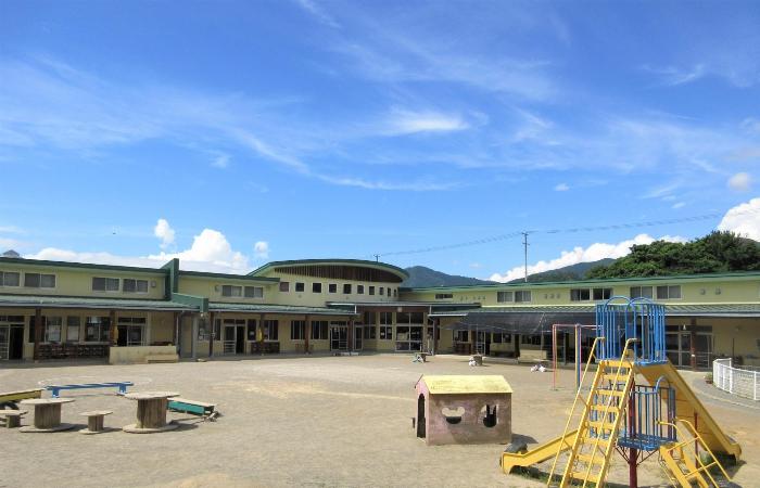 晴れた空を背景に薄黄色の外壁の1階建ての園舎があります。L字型の園舎の前には広い園庭が広がり、手前には鉄製の滑り台や上り棒、木の小屋、園児が作ったアスレチックなどがあります。