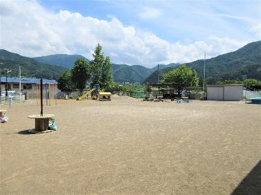 園舎から広い園庭を撮影した写真。青空のもと、奥には緑鮮やかな山が広がります。園庭の奥にある滑り台や上り棒が小さく見えます。
