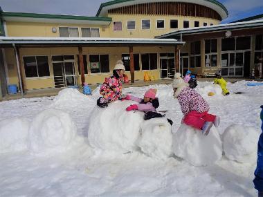 冬、一面に雪が積もった仁礼保育園の園庭に、大きな雪玉が6つ並んでいる。スキーウエアをきて帽子をかぶり手袋をはめた園児が、その雪玉の上にのって遊んでいる様子。