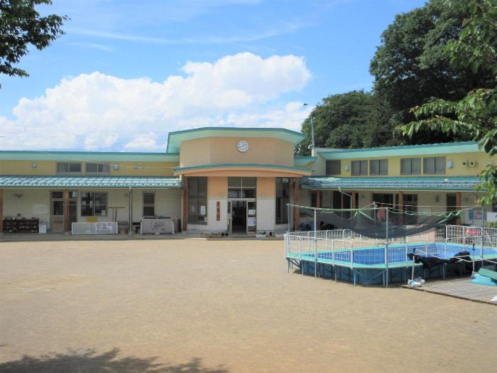 晴れた青空を背景にベージュ色の外壁の高甫保育園園舎の写真。1階建てで中央に玄関、L字型の園舎前には園庭が広がっています。。