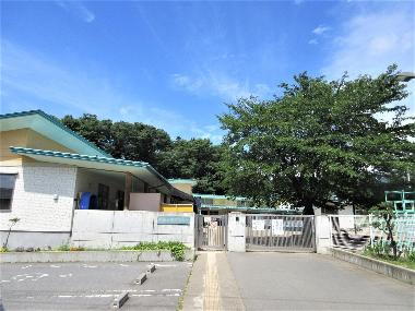 左手の園舎から続く玄関。白い壁と門からなり、インターフォンが設置されています。入って右側に桜の木がみえます。