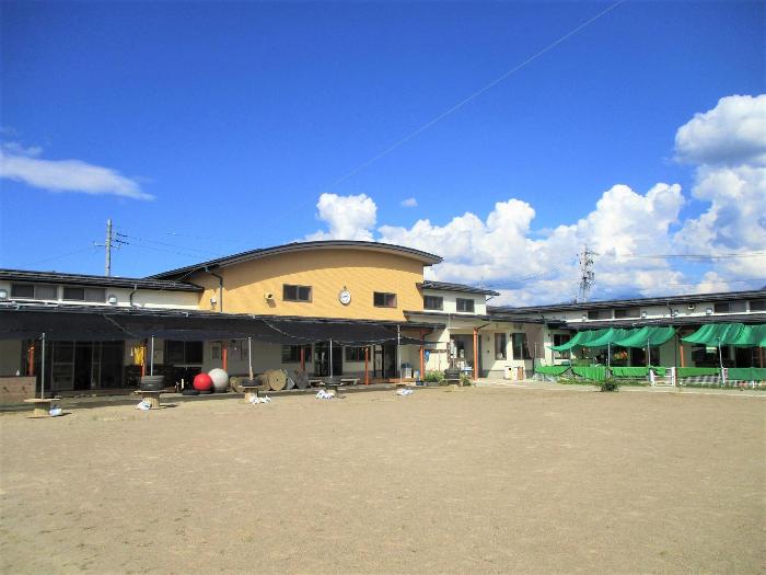 青空の下、ベージュと白色の外壁の須坂千曲保育園の園舎の写真。アーチ状の屋根。園舎の前には園庭が広がる。