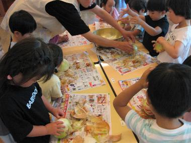 須坂東部保育園の保育室内で食育活動の一環として玉ねぎの皮をむいている園児の様子。保育士に教わりながら机の上で一人一つずつ玉ねぎの皮を丁寧にむいています。