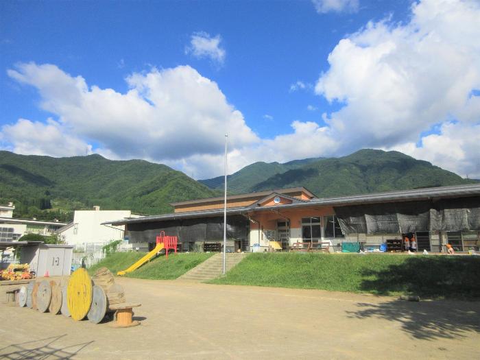 晴れた青空の下、奥には青々とした山並みが広がり、山のふもとに建つ豊丘保育園の1階建ての園舎の写真です。園舎から階段で10段ほど下がった場所に園庭が広がり、園庭中央には木のアスレチックがあります。園庭と園舎の間の傾斜は芝生になっています。