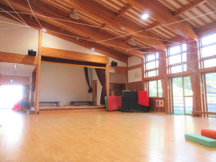 天井には木の梁があり、壁一面大きな窓で明るい日差しが入る豊丘保育園ホールの写真。奥にはステージ、ピアノがあります。
