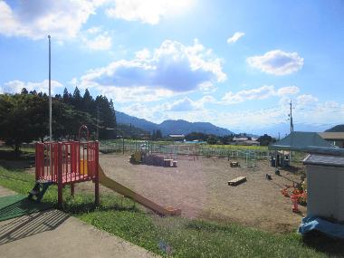 豊丘保育園園舎からの景色。手前にある園庭に降りる滑り台から園庭に続き、近くの林、遠くの山々と須坂市街を見下ろす景色が広がっています。