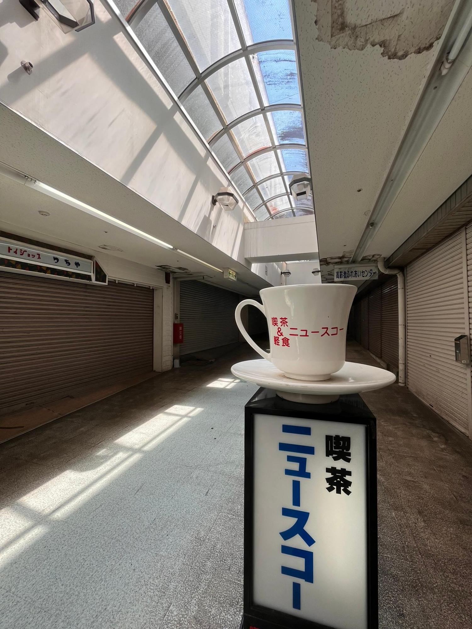 ショッピングセンター「パルム」で営業していた喫茶店「ニュースコー」の看板。「喫茶ニュースコー」と書かれ、看板上部にはコーヒーカップとソーサーの置物が乗っている。他の店舗はすべてシャッターを下ろしている。人通りもない。