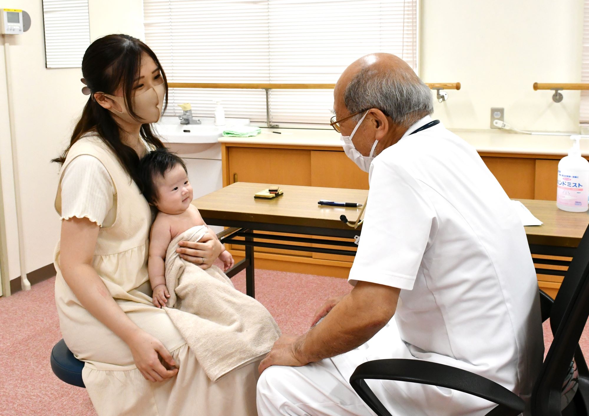 乳幼児を抱いた女性がと白衣を着た年配の医師が向かい合って座っている。こちらを向いている乳幼児と女性の顔、医師の横顔が見え、いずれも笑顔を浮かべている。