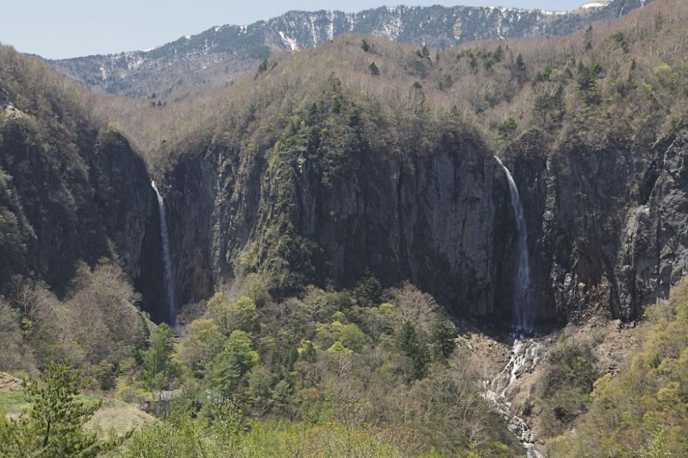 雪の残る山々を背景に、深く切れ込んだ暗い岩肌の崖から二筋の滝が勢いよく流れ落ち、その下には豊かな緑の木々が生い茂り、急流となって合流する壮大な自然の風景の写真