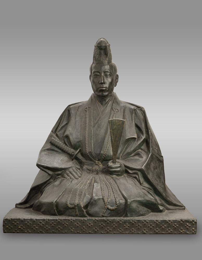 束帯姿で烏帽子を被り、太刀を腰に差し、扇を手に持って座っている堀直虎公の姿を再現した銅像の写真
