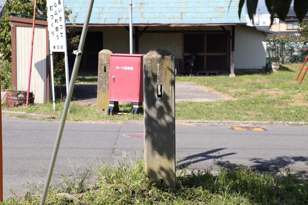 芝と雑草が生えた道路の脇に、日吉社（日吉山王）跡地を示す文字が刻まれた石柱が手前にあり、背景には赤いホース格納箱と板張りの建物が見える、日吉社跡地を写した写真