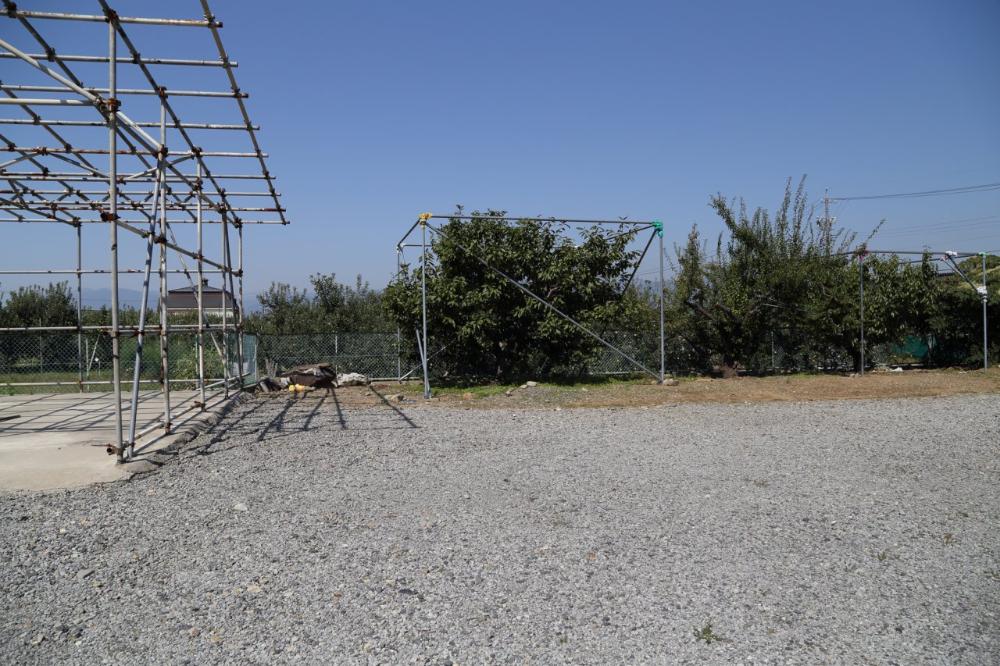手前は砂利敷きの更地、左側には足場の鉄骨、中央奥にはフェンスと植栽が見え、周囲は畑や住宅に囲まれた開放的な神明社(神明宮)跡地の写真